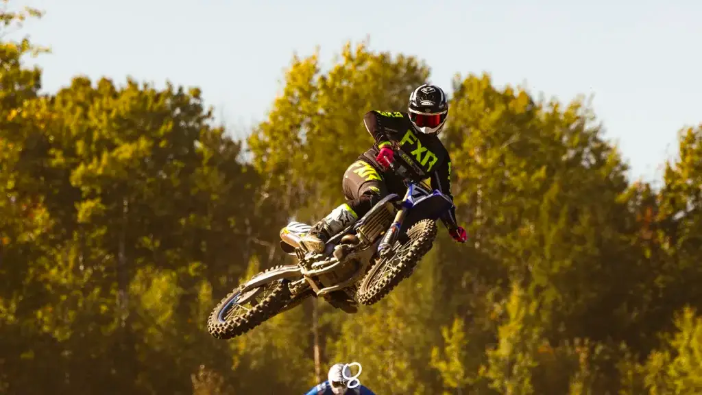Un pilote de motocross en tenue noire et verte effectue un saut en l'air sur une moto de terre sur fond d'arbres et de ciel dégagé.