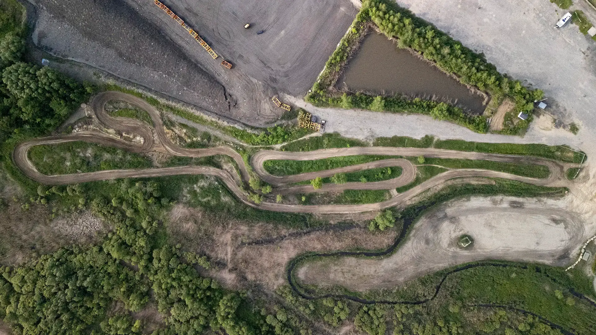 Vue aérienne d'une piste de course en terre sinueuse entourée de verdure, d'arbres et de quelques zones de gravier, avec un étang et des véhicules visibles dans le coin supérieur droit.
