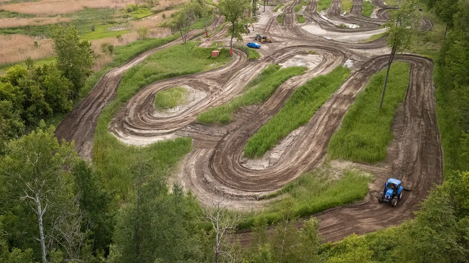 Vue aérienne d'une piste de course sinueuse en terre battue, entourée d'arbres et d'herbe, avec un tracteur bleu garé sur la piste dans le coin inférieur droit.