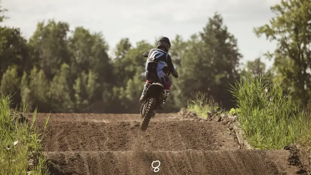 Un pilote de motocross portant un casque et un équipement accélère sur une piste de terre entourée d'arbres verts et de hautes herbes par une journée ensoleillée. De la poussière s'échappe de la roue arrière.