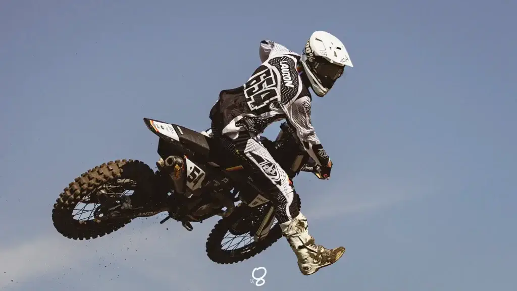 Un pilote de motocross en tenue complète effectue un saut sur une moto tout-terrain dans un ciel bleu clair, la terre s'envolant du pneu arrière.