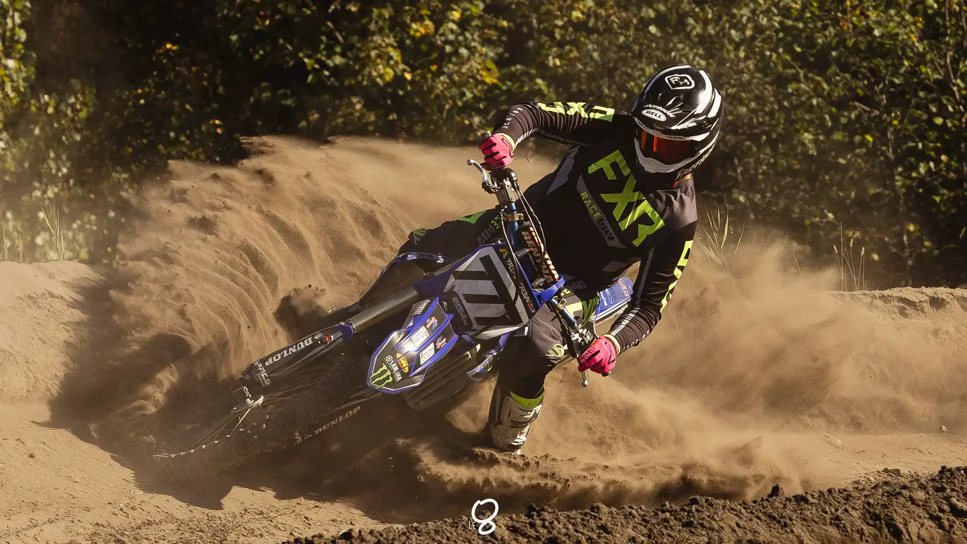 Un pilote de motocross en tenue complète, comprenant un casque et des gants, prend un virage serré sur une piste en terre battue, soulevant un nuage de poussière avec sa moto. Des arbres et du feuillage sont visibles à l'arrière-plan.