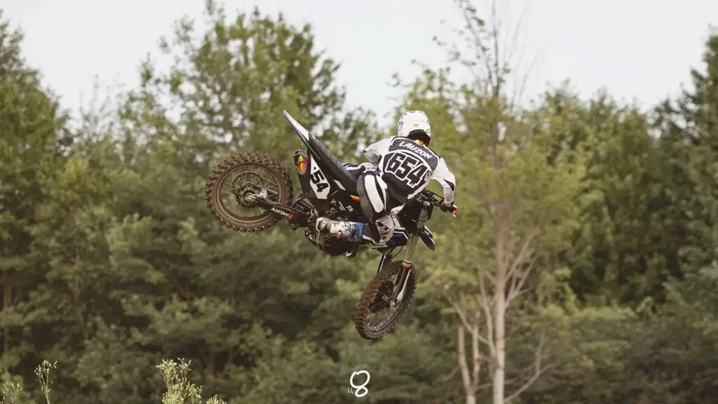 Un pilote de motocross en tenue noire et blanche, numéroté 654, effectue une acrobatie aérienne sur une moto tout-terrain sur fond d'arbres verts.