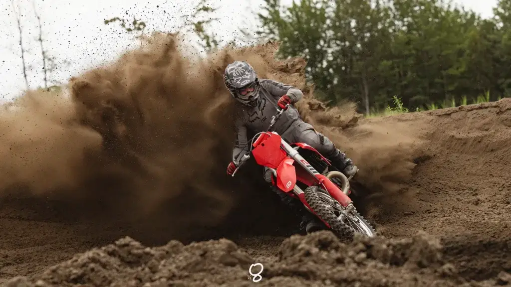 Une personne en tenue de motocross conduit une moto de terre rouge dans de la terre meuble, créant une spectaculaire gerbe de terre derrière elle. Des arbres et un ciel nuageux sont visibles à l'arrière-plan.