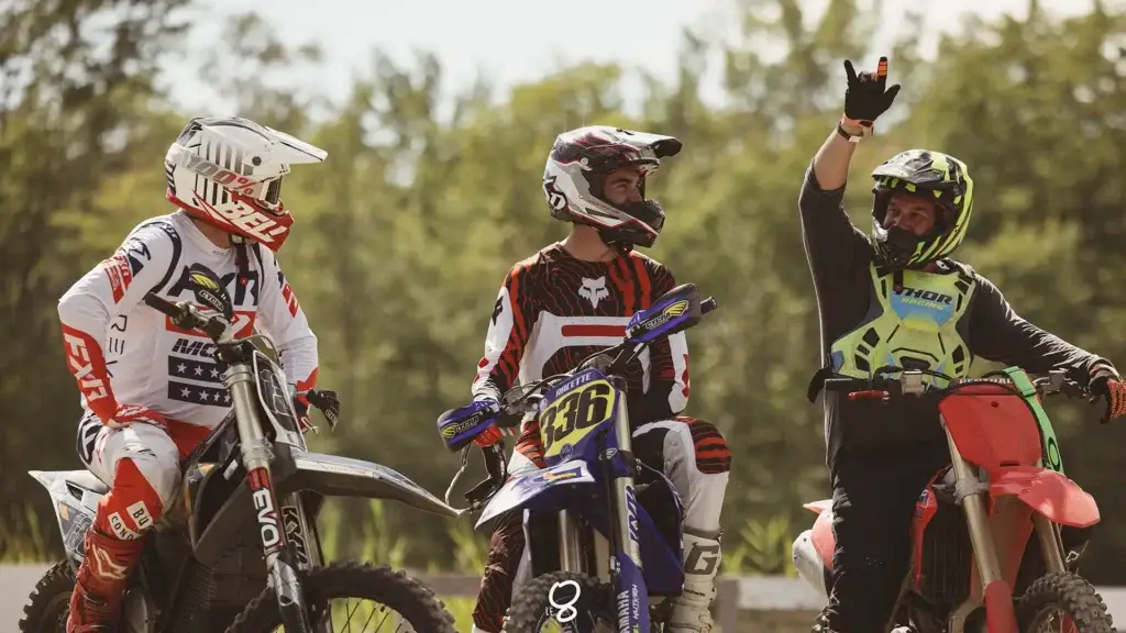 Trois pilotes de motocross en tenue colorée sont assis sur leurs motos en plein air. L'un d'eux, à droite, vêtu d'un gilet vert, montre le ciel tandis que les autres regardent et sourient, avec des arbres et de la verdure en arrière-plan.