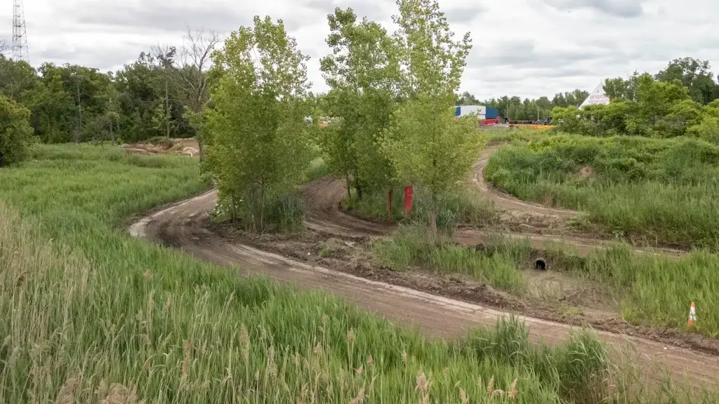 Un chemin de terre boueux serpente à travers de hautes herbes vertes et des arbres dans une zone rurale, avec un ciel nuageux au-dessus de la tête et quelques bâtiments visibles au loin.