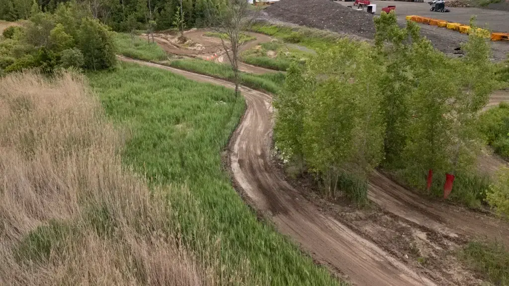 Un chemin de terre sinueux traverse une zone verte et herbeuse, bordée de part et d'autre de grands arbres et de buissons. À l'arrière-plan, des conteneurs et des équipements industriels sont visibles près d'un terrain dégagé.