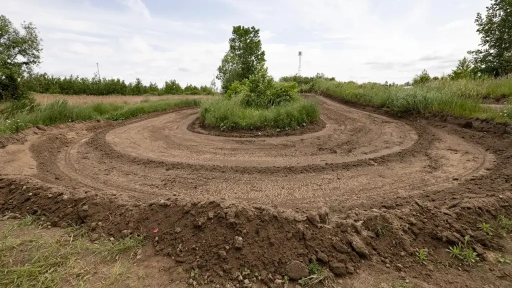 Un chemin de terre s'enroule autour d'une île herbeuse avec des arbres et de hautes herbes au centre, entourée d'autres herbes et d'un ciel dégagé. La piste semble avoir été faite récemment, avec des marques de pneus visibles dans le sol.