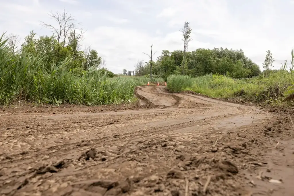 Un chemin de terre boueux serpente à travers l'herbe verte et les arbres sous un ciel nuageux, avec des cônes de signalisation orange visibles au loin.
