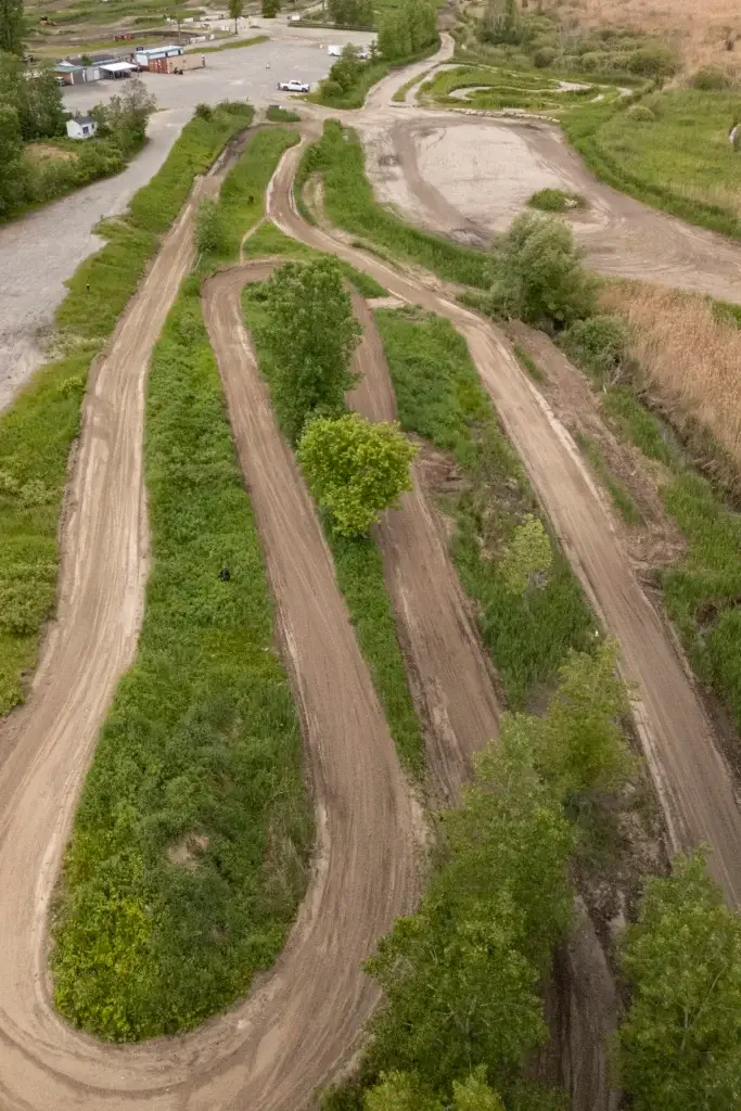Vue aérienne d'un chemin de terre sinueux avec de multiples courbes, entouré d'herbe verte et d'arbres épars, à côté d'une zone ouverte avec des véhicules garés et des bâtiments.