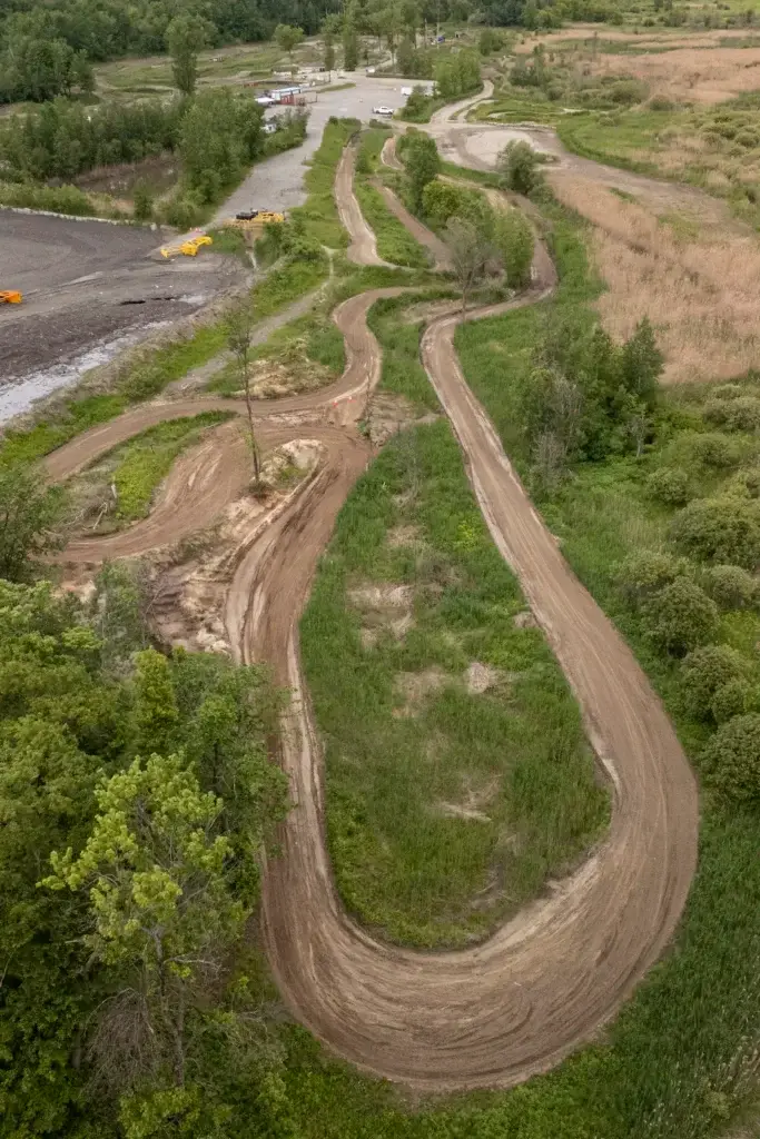 Un chemin de terre sinueux traverse une végétation verdoyante et des champs herbeux, avec des arbres et des buissons de part et d'autre. À l'arrière-plan se trouve une aire de stationnement avec plusieurs véhicules et équipements.