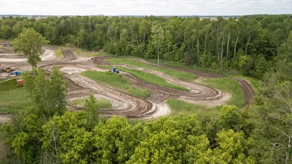 Vue aérienne d'une piste de motocross serpentant à travers des zones herbeuses et entourée d'une forêt verte dense sous un ciel partiellement nuageux.