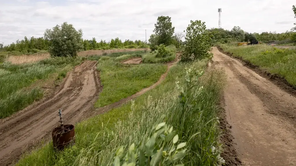 Sous un ciel nuageux, une piste de motocross aux tracés sinueux et boueux serpente à travers des champs herbeux et des arbres épars. Un vieux tonneau en métal se trouve près de la piste, et de hautes herbes entourent la zone.