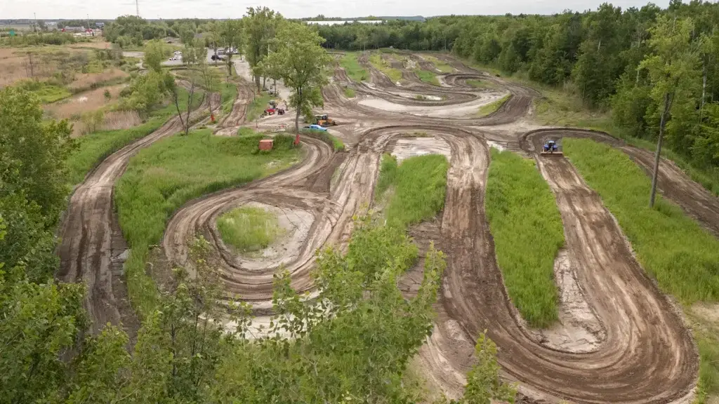 Une piste de motocross sinueuse avec de multiples virages et des zones vertes et herbeuses traverse un paysage boisé, vue d'en haut par une journée nuageuse.
