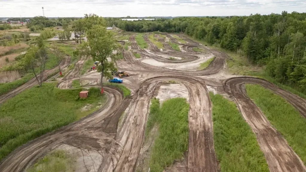Vue aérienne d'une piste de course sinueuse en terre battue, entourée d'herbe verte et d'arbres, avec quelques véhicules et équipements garés près de la piste. Le ciel est nuageux et la région est essentiellement rurale.