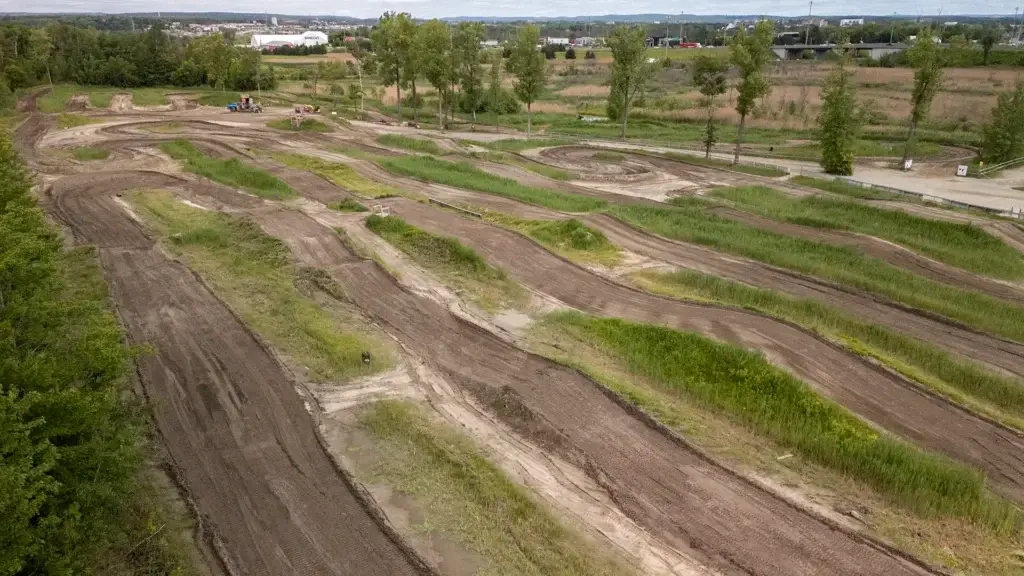 Vue aérienne d'une piste de motocross en terre battue avec des chemins sinueux et des sections herbeuses, entourée d'arbres et de champs, sous un ciel nuageux.