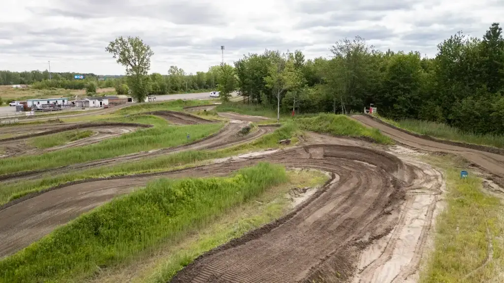 Une piste de motocross sinueuse avec de nombreux virages et des zones herbeuses, entourée d'arbres verts et d'un ciel nuageux. Quelques véhicules et bâtiments sont visibles à l'arrière-plan, près d'une route.