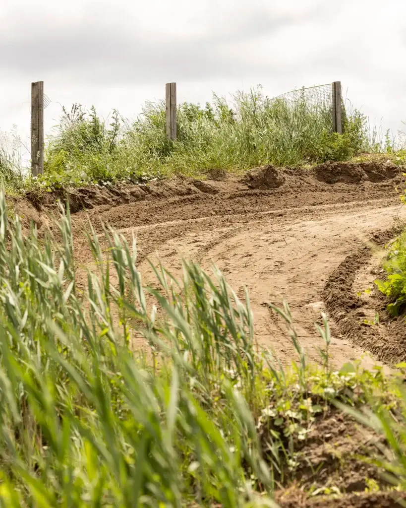 Un chemin de terre avec des traces de pneus serpente à travers les hautes herbes vertes, bordé de poteaux et de fils de clôture en bois, sous un ciel nuageux.