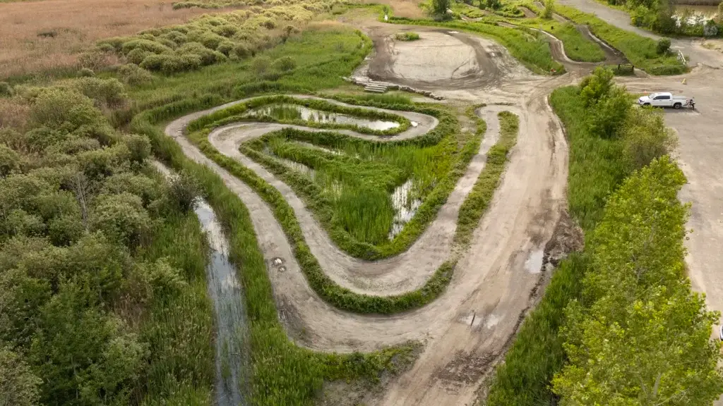 Vue aérienne d'un chemin de terre sinueux entouré de végétation verte, avec des points d'eau et une petite aire de stationnement avec un véhicule blanc visible dans le coin supérieur droit.