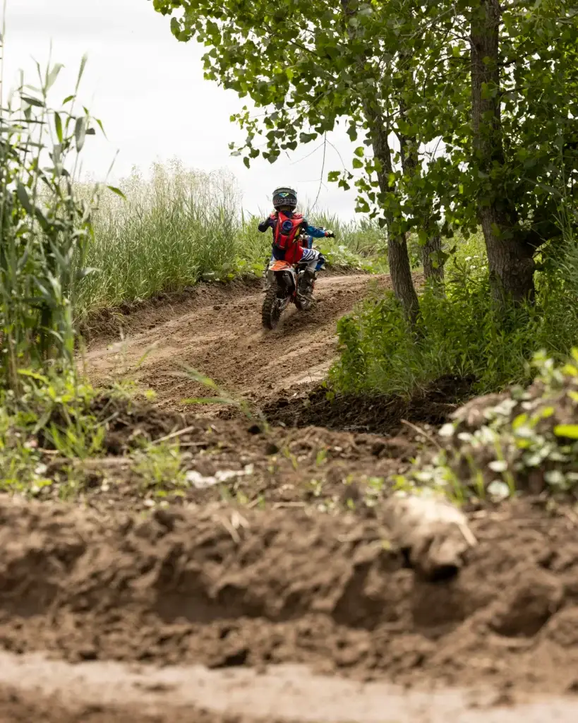 Un enfant portant un casque et une tenue de motocross conduit une petite moto sur une piste sinueuse entourée d'herbes hautes et d'arbres.