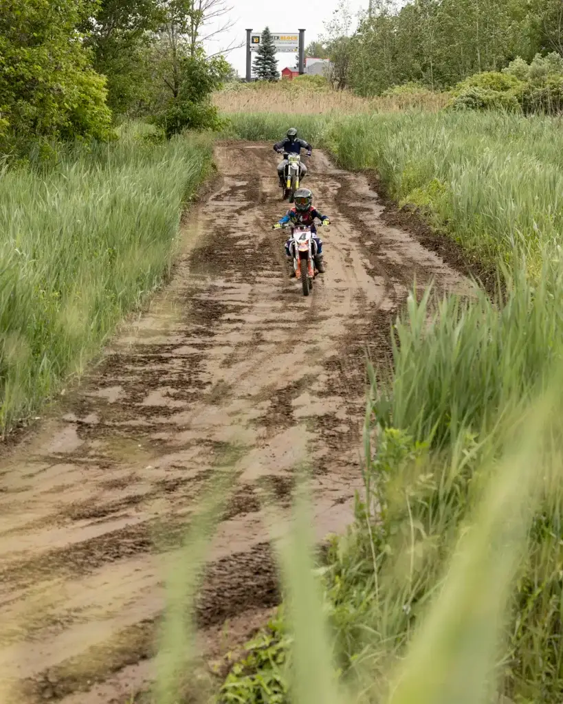 Deux personnes conduisent des motos tout-terrain sur un sentier boueux entouré de hautes herbes vertes et d'arbres. Un autre motard est visible à l'arrière-plan, près d'un panneau.