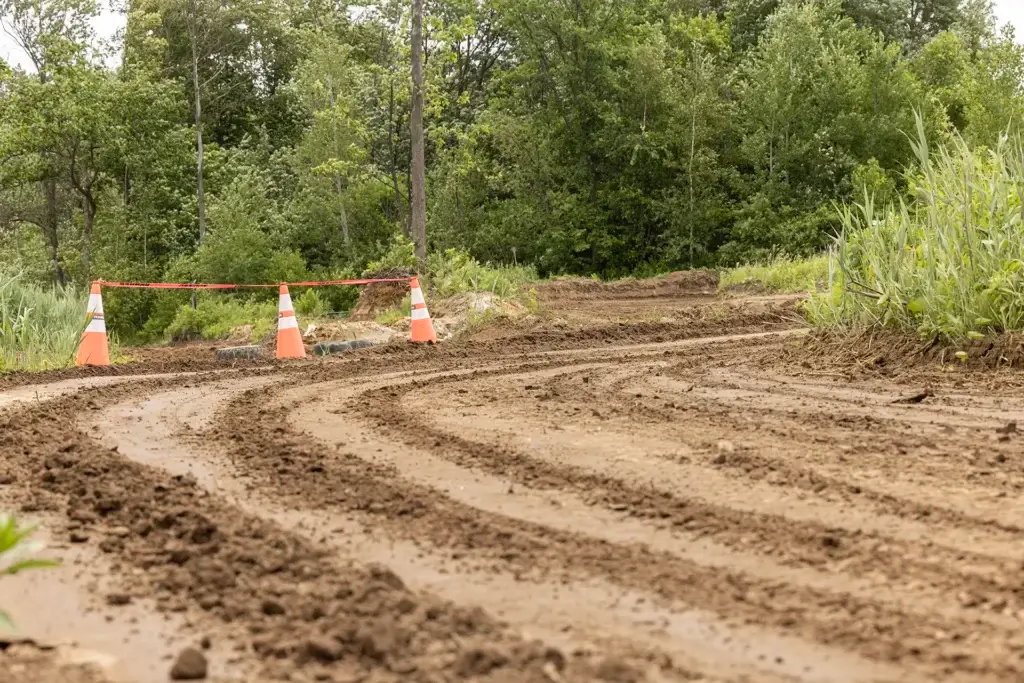 Un chemin de terre boueux traverse une zone verte et boisée. Trois cônes de signalisation orange et du ruban rouge bloquent une partie du chemin, indiquant une fermeture ou un danger à venir.