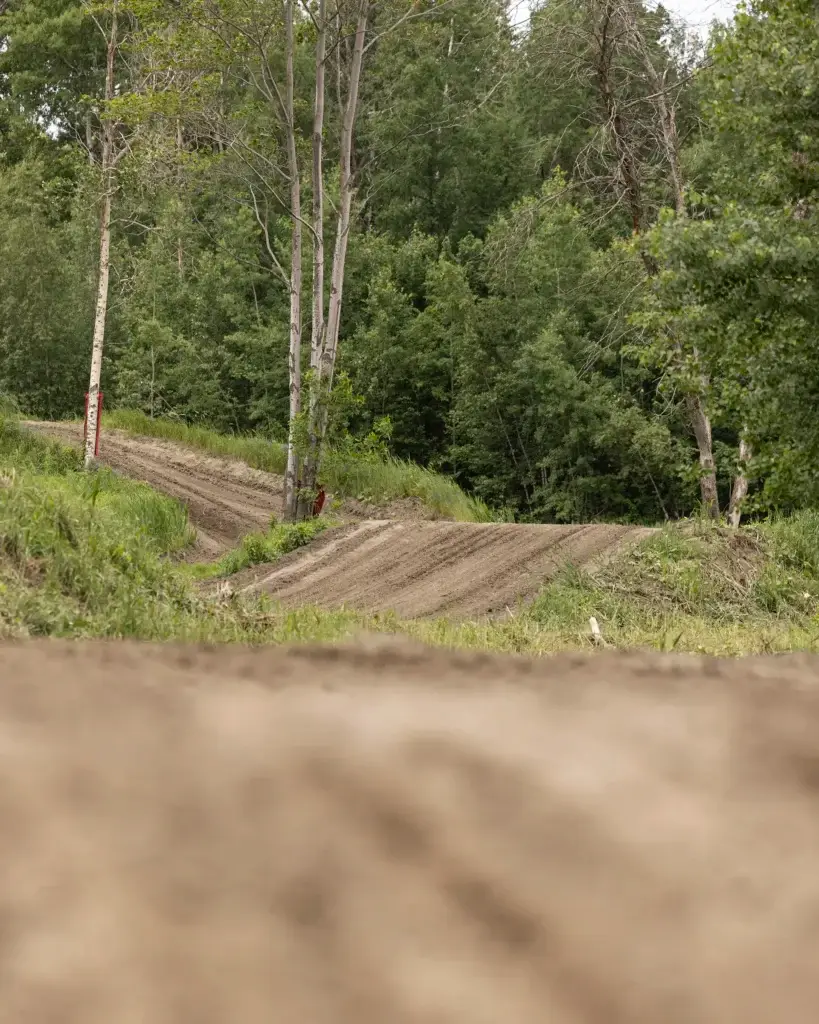 Une piste de motocross traverse une zone herbeuse et boisée avec des arbres verts en arrière-plan. La piste est vide et le premier plan montre une section floue de terre.