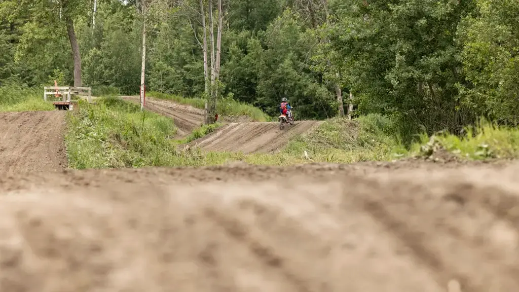 Une personne conduit une moto tout-terrain sur une piste de motocross sinueuse, entourée d'arbres et d'herbe verdoyants. Le premier plan montre la surface rugueuse et incurvée de la piste, tandis que l'arrière-plan est rempli d'une forêt dense.