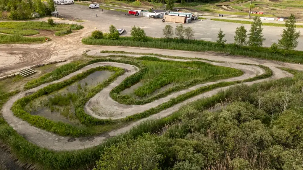 Un chemin de terre sinueux entouré de verdure et d'eau, avec un parking, des arbres, des véhicules et un petit bâtiment en arrière-plan, sous un ciel partiellement nuageux.