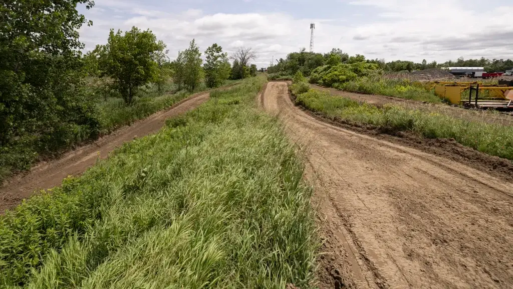 Un chemin de terre serpente à travers la végétation herbeuse et les arbres sous un ciel partiellement nuageux. Des engins de chantier sont visibles au loin sur le côté droit.
