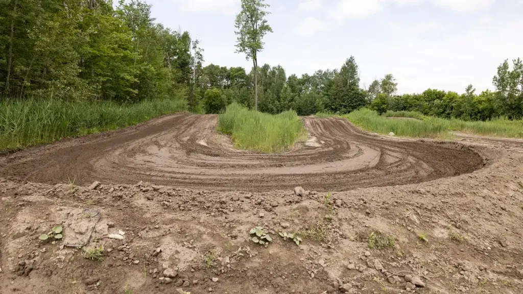 Une piste de motocross serpente à travers l'herbe verte et les arbres sous un ciel partiellement nuageux, les traces de pneus étant visibles dans le sol meuble.