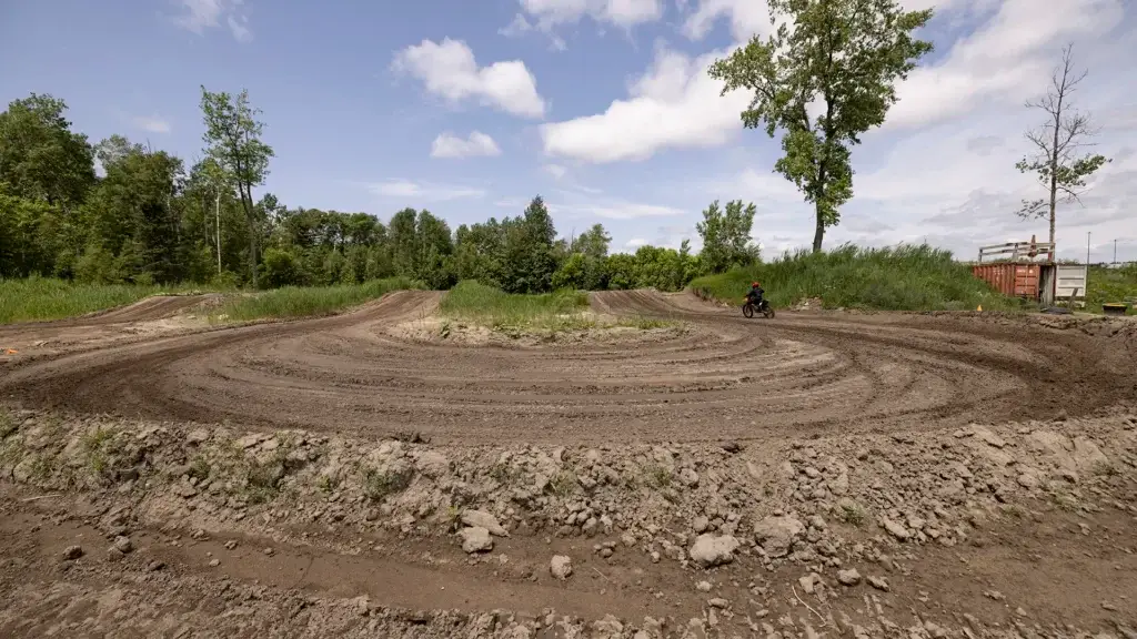 Une piste de motocross traverse une zone herbeuse parsemée d'arbres et de buissons. Un motocycliste seul est visible au centre droit. Le ciel est partiellement nuageux et le soleil brille.