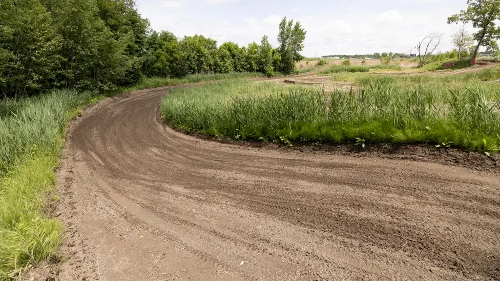 Un large chemin de terre s'incurve vers la gauche à travers un terrain herbeux, bordé de grandes plantes vertes et d'arbres, sous un ciel partiellement nuageux. Le chemin semble être destiné aux véhicules tout-terrain ou aux motocross.