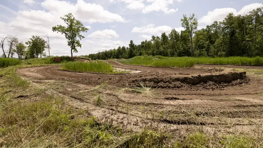 Une piste de motocross serpente à travers des champs herbeux et des arbres sous un ciel partiellement nuageux, avec des traces de pneus visibles et des taches de boue sur le sol.