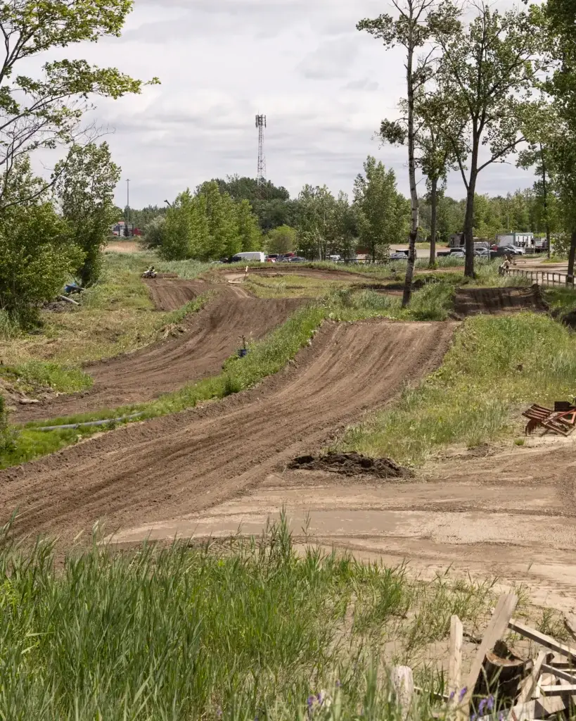 Sous un ciel nuageux, une piste de motocross serpente à travers une zone herbeuse et bordée d'arbres, avec des voitures garées et quelques bâtiments visibles à l'arrière-plan.