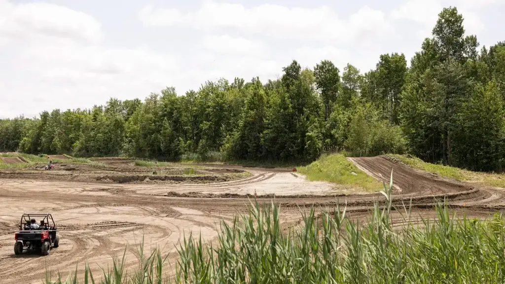 Sous un ciel partiellement nuageux, une piste de course en terre battue, pratiquement vide et marquée de traces de pneus, traverse une zone ouverte bordée de hautes herbes vertes et d'arbres touffus. Un petit véhicule tout-terrain est garé sur le côté gauche de la piste.