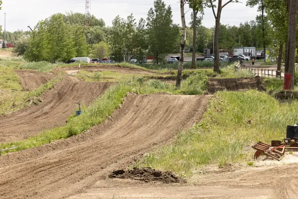 Une piste de motocross en terre battue avec des virages sinueux et surélevés et des zones herbeuses, entourée d'arbres. Plusieurs véhicules sont garés à l'arrière-plan, près d'une rangée d'arbres et d'une clôture.