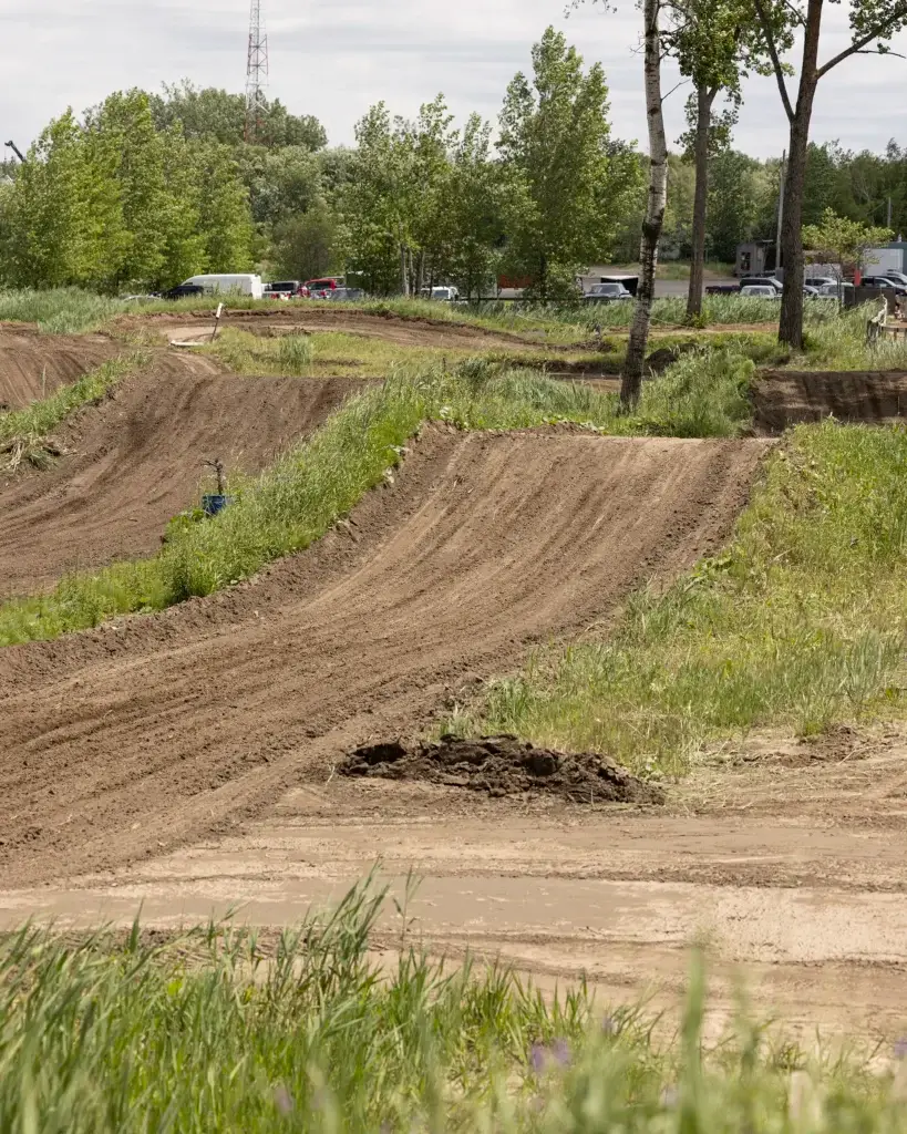 Une piste de motocross avec des chemins sinueux et des sauts, entourée d'herbe verte et d'arbres. Plusieurs véhicules sont garés à l'arrière-plan sous un ciel nuageux.