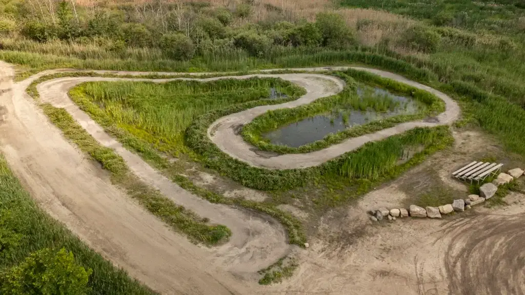 Vue aérienne d'un chemin de terre sinueux formant un cœur autour d'un petit étang, entouré d'herbe, de buissons et de pierres éparses. Un banc en bois se trouve près du chemin sur le côté droit.