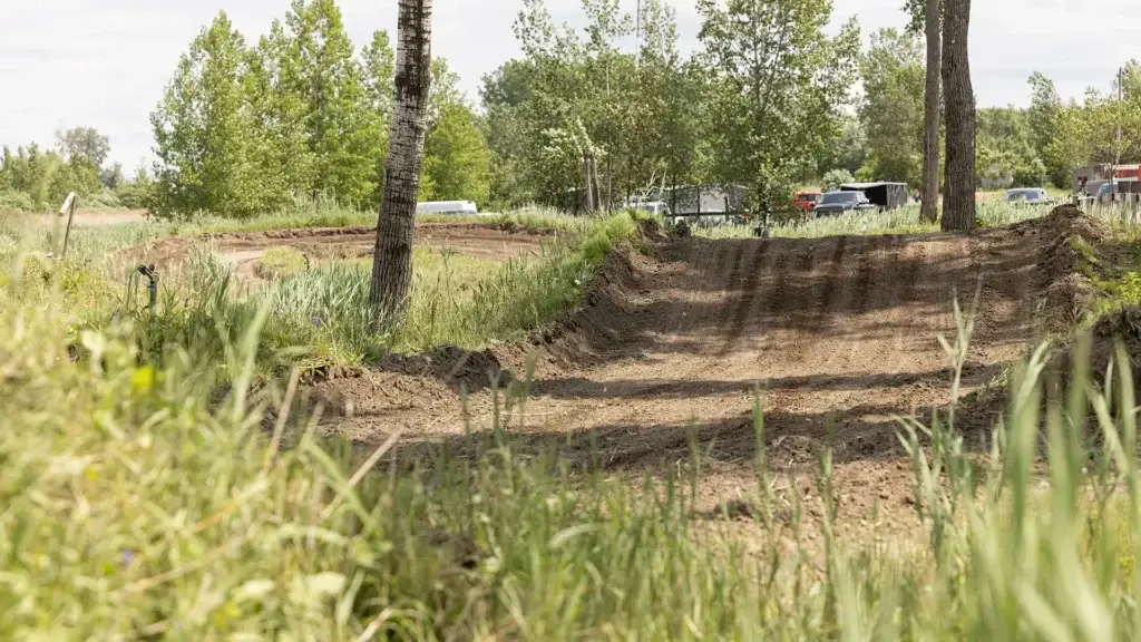 Une piste de motocross serpente sur un terrain herbeux, entouré d'arbres verts. À l'arrière-plan, des véhicules et une clôture sont partiellement visibles sous un ciel clair et partiellement nuageux.