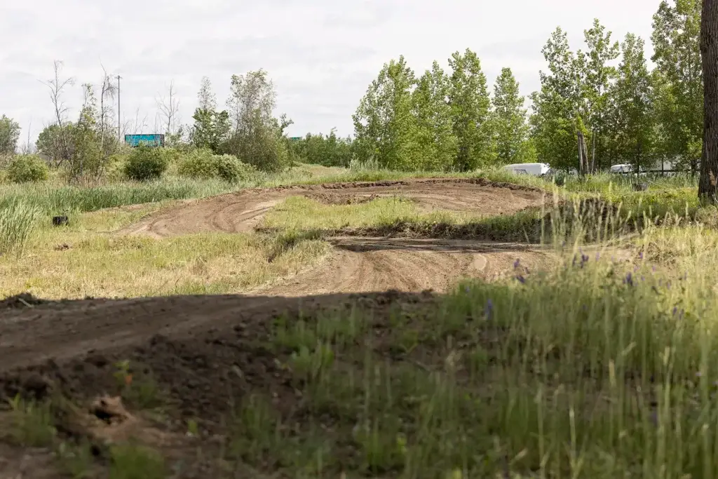 Une piste de motocross traverse un terrain herbeux avec des arbres et des buissons en arrière-plan, sous un ciel nuageux. La piste est vide et entourée de végétation verte.