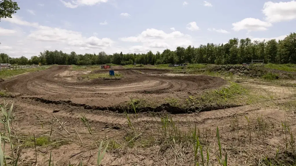 Une piste de motocross en terre battue, avec des traces de pneus incurvées et boueuses, serpente à travers un terrain herbeux, entouré d'arbres verts et d'un ciel partiellement nuageux.