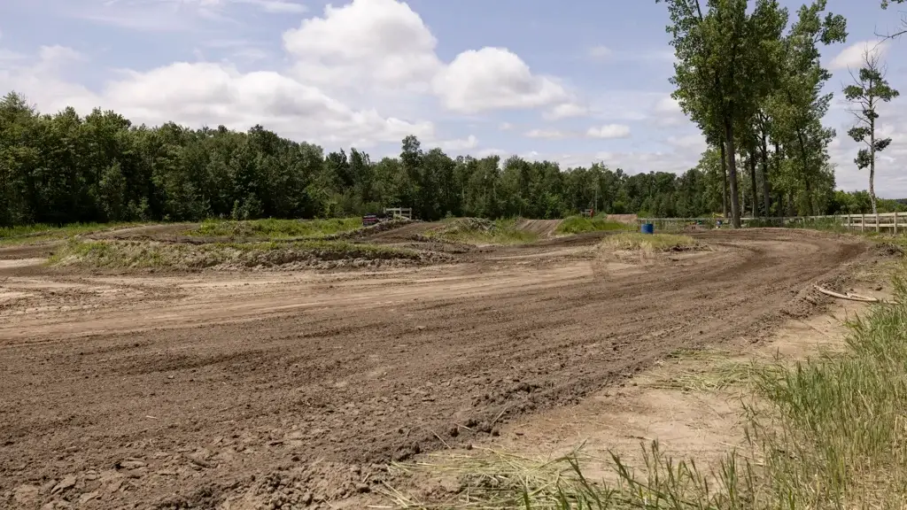 Une piste de motocross traverse une zone herbeuse et dégagée, entourée d'arbres verts denses, sous un ciel partiellement nuageux. La piste semble vide et le sol est sec et rugueux.