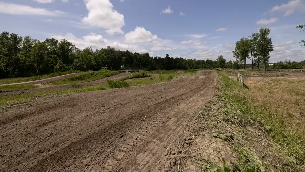 Une piste de motocross en terre battue avec des virages sinueux, entourée d'herbe verte, d'arbres et d'un ciel bleu partiellement nuageux. La zone semble vide, aucun pilote n'étant présent.