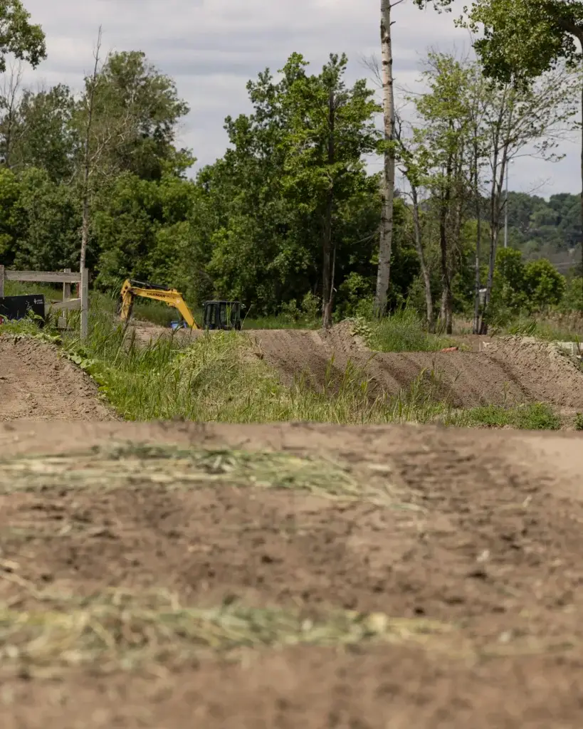 Une pelleteuse jaune est assise près de monticules de terre entourés d'herbe verte et d'arbres sous un ciel nuageux, suggérant des travaux de construction ou d'aménagement paysager dans une zone rurale.