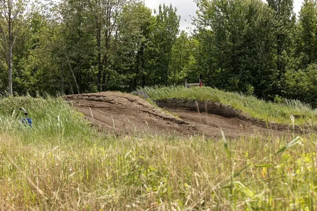 Une piste de dirt bike avec de hautes bermes est entourée d'herbes hautes et d'arbres verts denses sous un ciel partiellement nuageux.