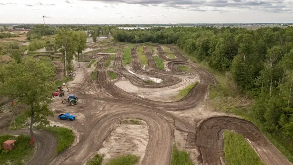 Vue aérienne d'une piste de motocross sinueuse entourée d'arbres verts et d'herbe, avec quelques véhicules et machines garés près de la piste sous un ciel nuageux.