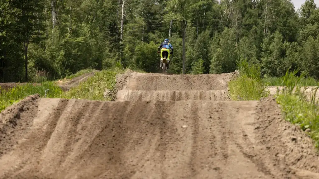 Une personne conduit une moto tout-terrain sur une piste de motocross entourée d'arbres verts, en s'éloignant de la caméra et en se dirigeant vers une série de sauts sur le chemin de terre.