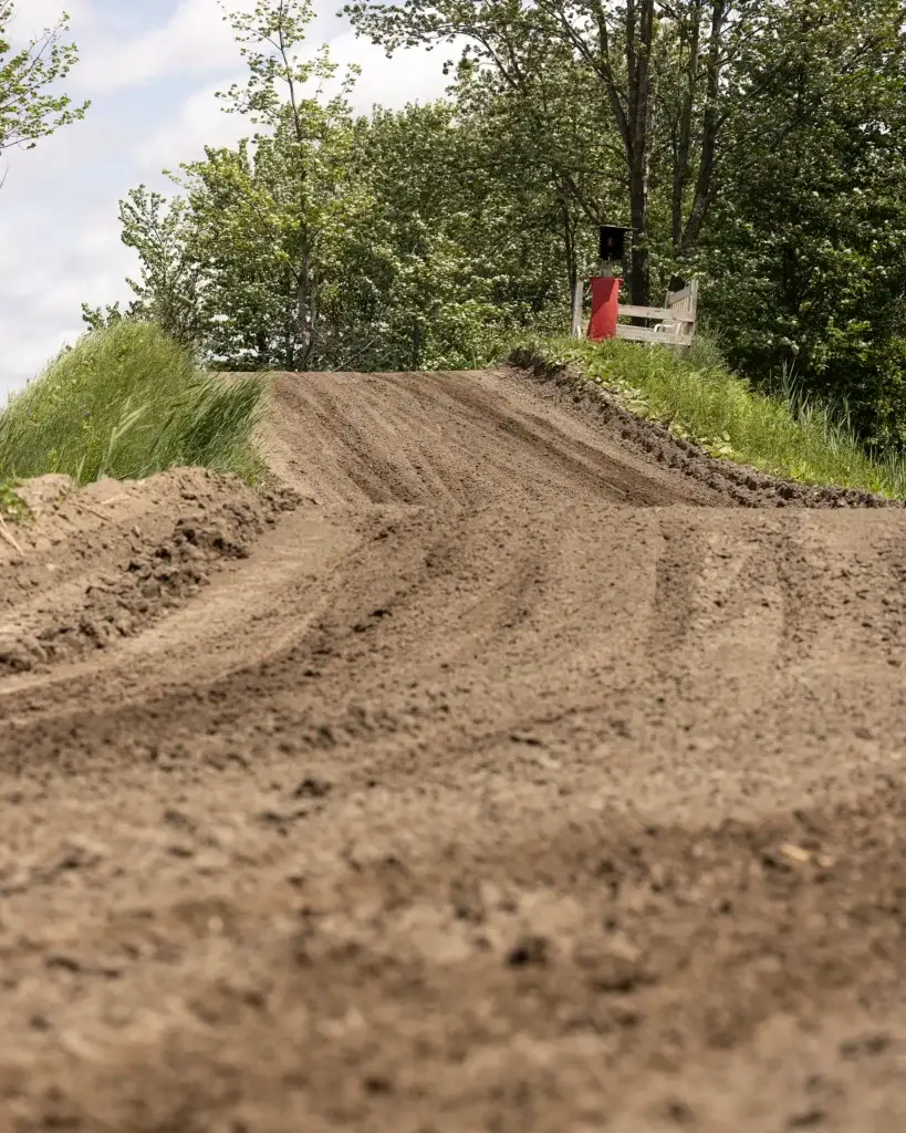 Une piste de course en terre montée, bordée d'herbe et d'arbres. À l'arrière-plan, une tribune en bois blanc avec un signal rouge et noir se trouve près du bord de la piste, sous un ciel partiellement nuageux.