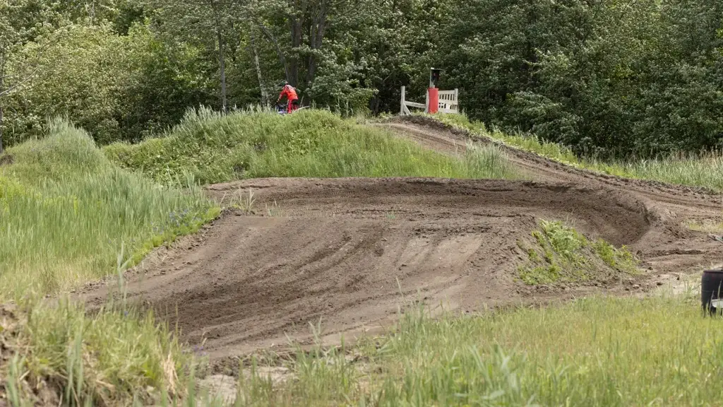 Une piste de motocross en terre battue avec des traces de pneus et de l'herbe verte des deux côtés, entourée d'arbres. Une personne en chemise rouge se tient près d'une structure en bois à l'arrière-plan.